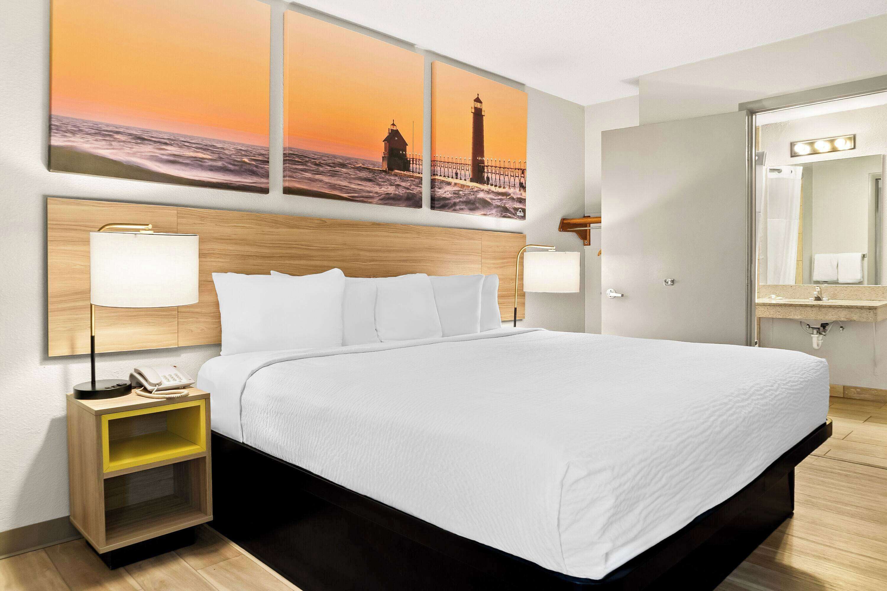 Habitació d'hotel amb un llit gran, dues taules de nit amb làmpades, un taulell amb un telèfon, un mirall amb il·luminació i una paret amb tres quadres de paisatge al capvespre.
