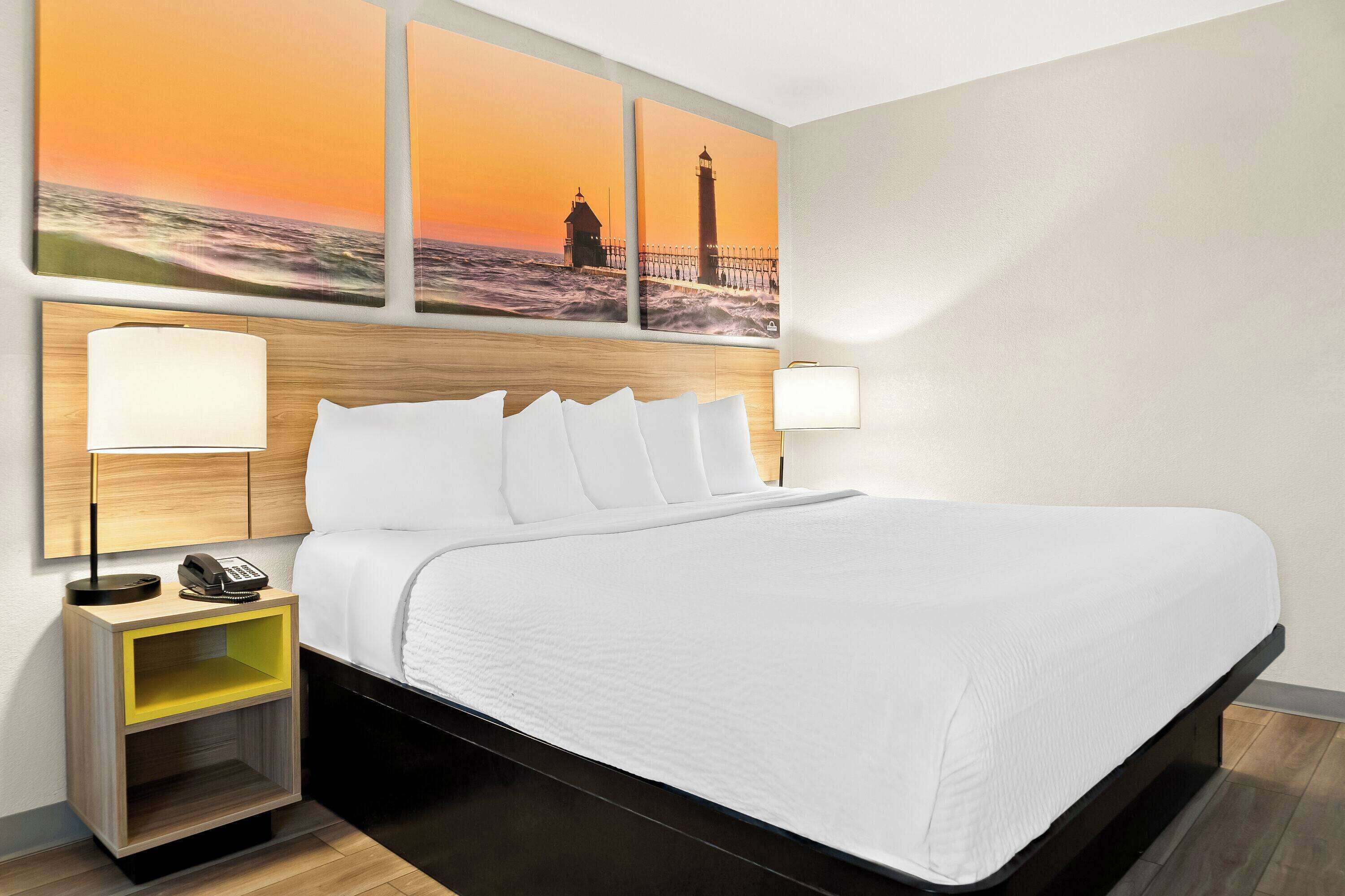 A hotel room with a bed, two bedside lamps, a nightstand with a phone, and a wall with three sunset paintings.