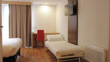 A hotel room with two single beds, a desk with a chair, a television mounted on the wall, and a radiator.