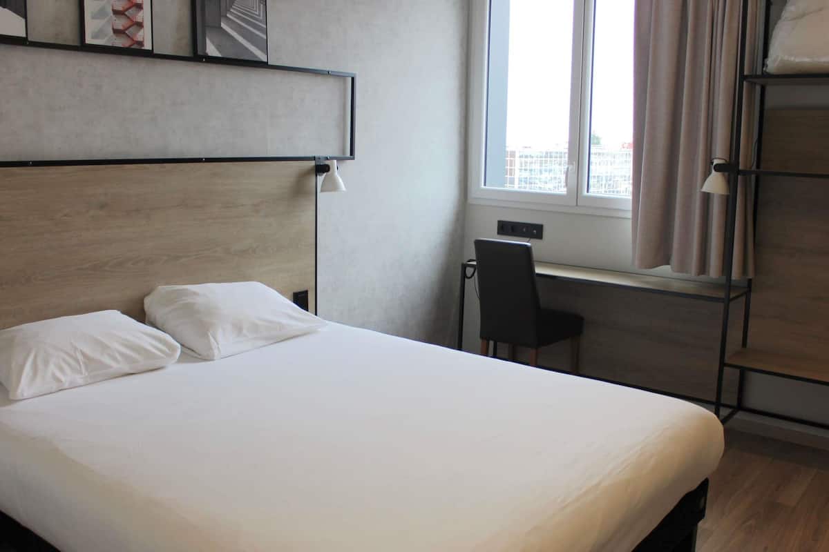 Una habitación de hotel con cama, escritorio, silla y ventana con cortinas.