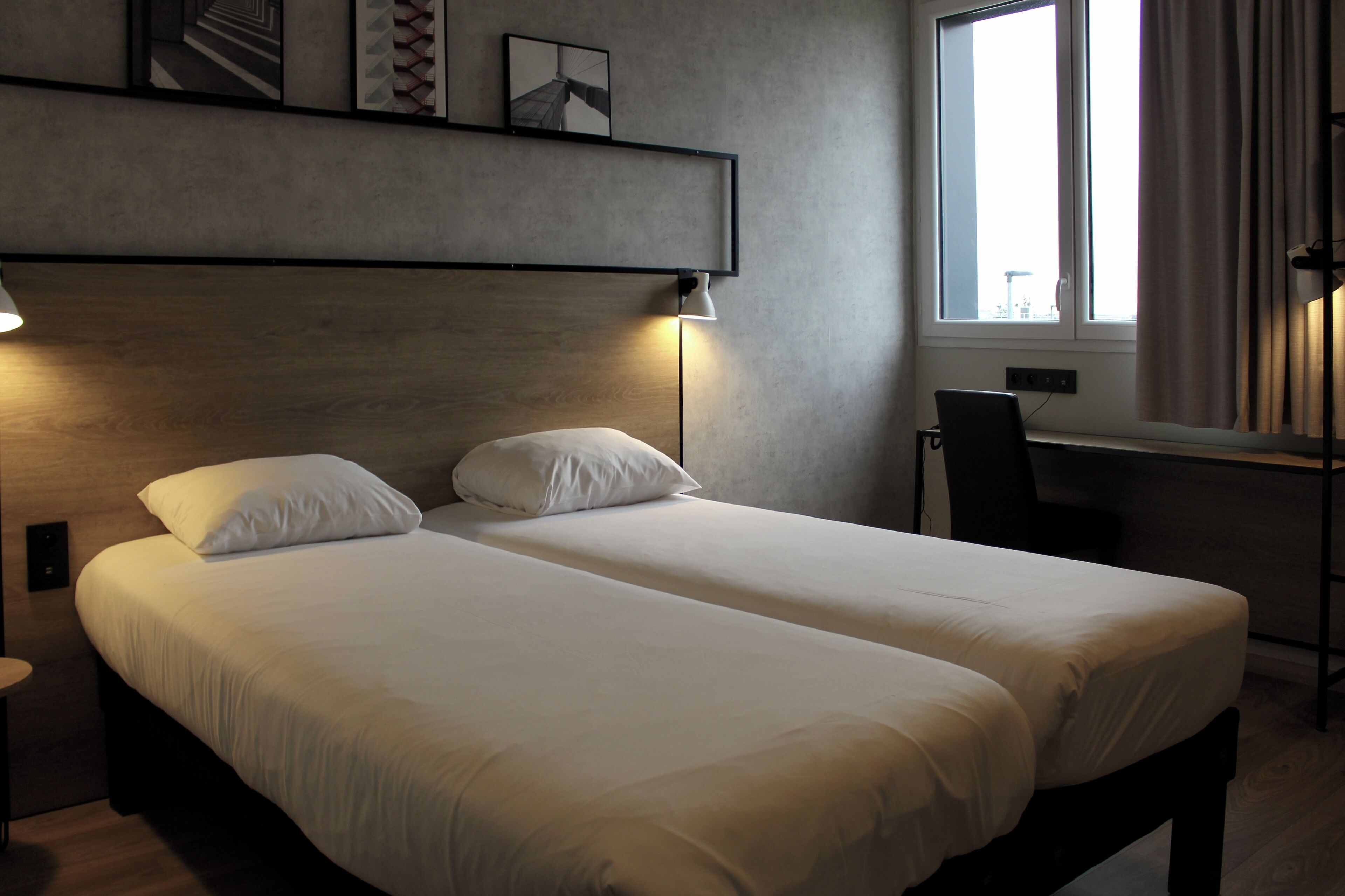 A double bed with white linens, a bedside table, a lamp, a chair, and a window with curtains.