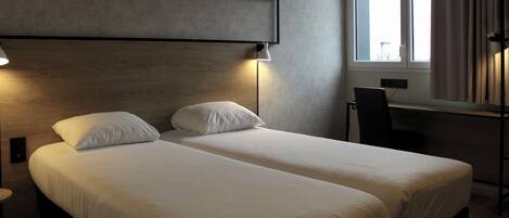 A double bed with white linens, a bedside table, a lamp, a chair, and a window with curtains.