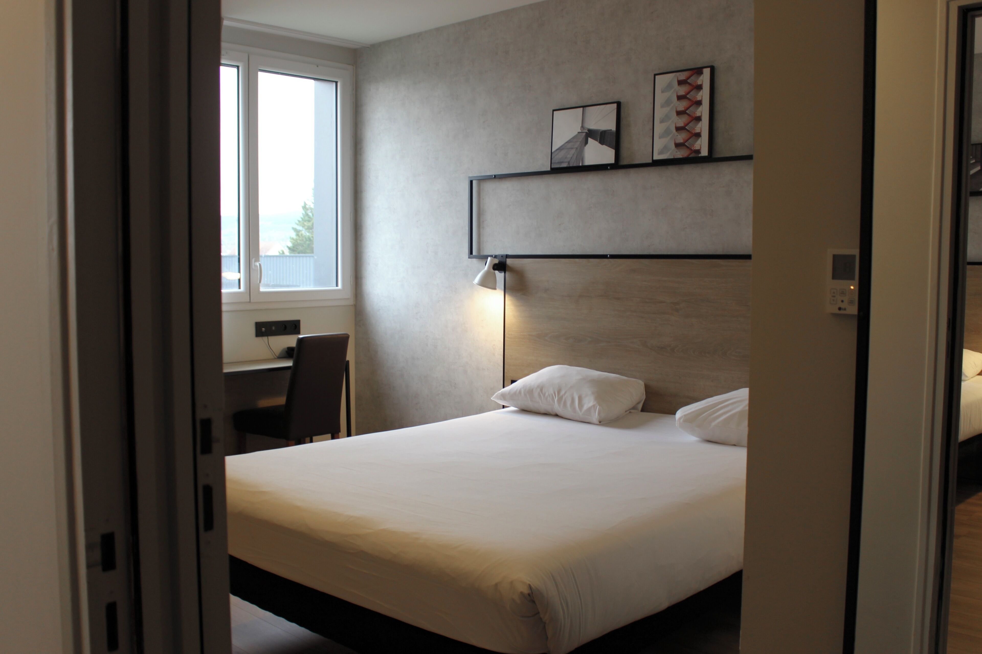 Une chambre d’hôtel avec un grand lit, un bureau avec une chaise, une fenêtre et une étagère fixée au mur, ornée de tableaux.