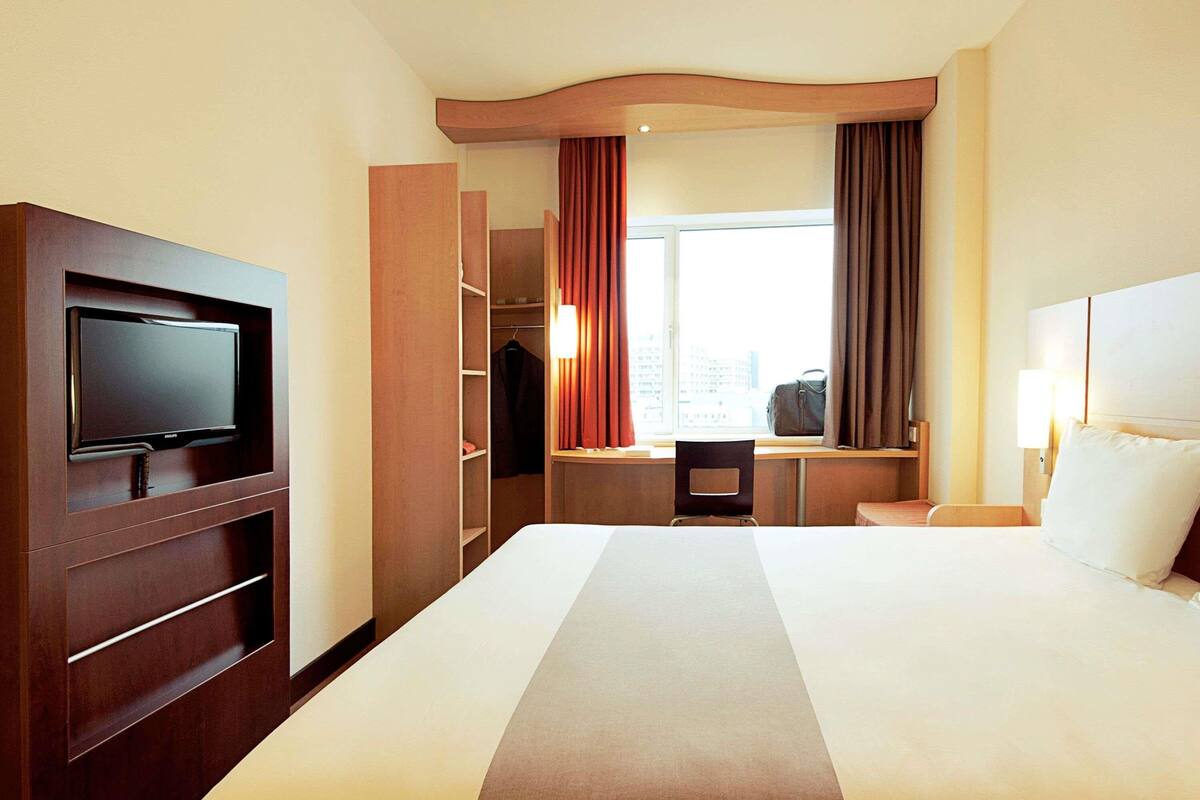Una habitación de hotel con cama, televisor montado en un mueble de madera, escritorio con silla y ventana con cortinas.