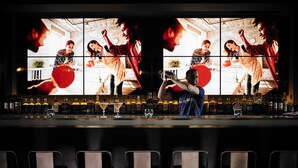 A bar area with multiple screens displaying people dancing, a bartender, and a variety of bottles and glasses on the bar counter.