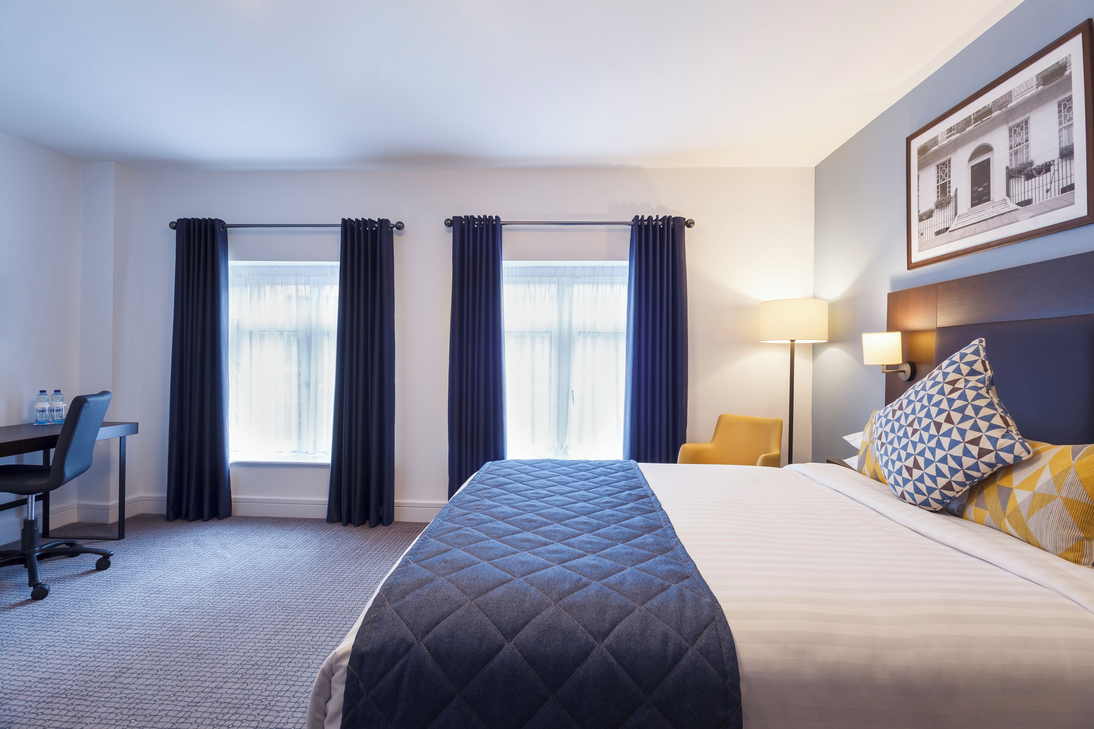 Una habitación de hotel con una cama, un escritorio, dos ventanas con cortinas, una lámpara y un cuadro en la pared.