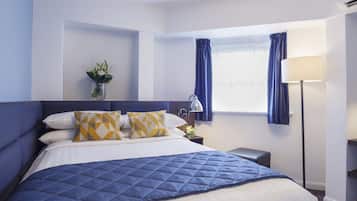A neatly made bed with a blue quilt, a bedside lamp, and a vase with flowers on the nightstand.