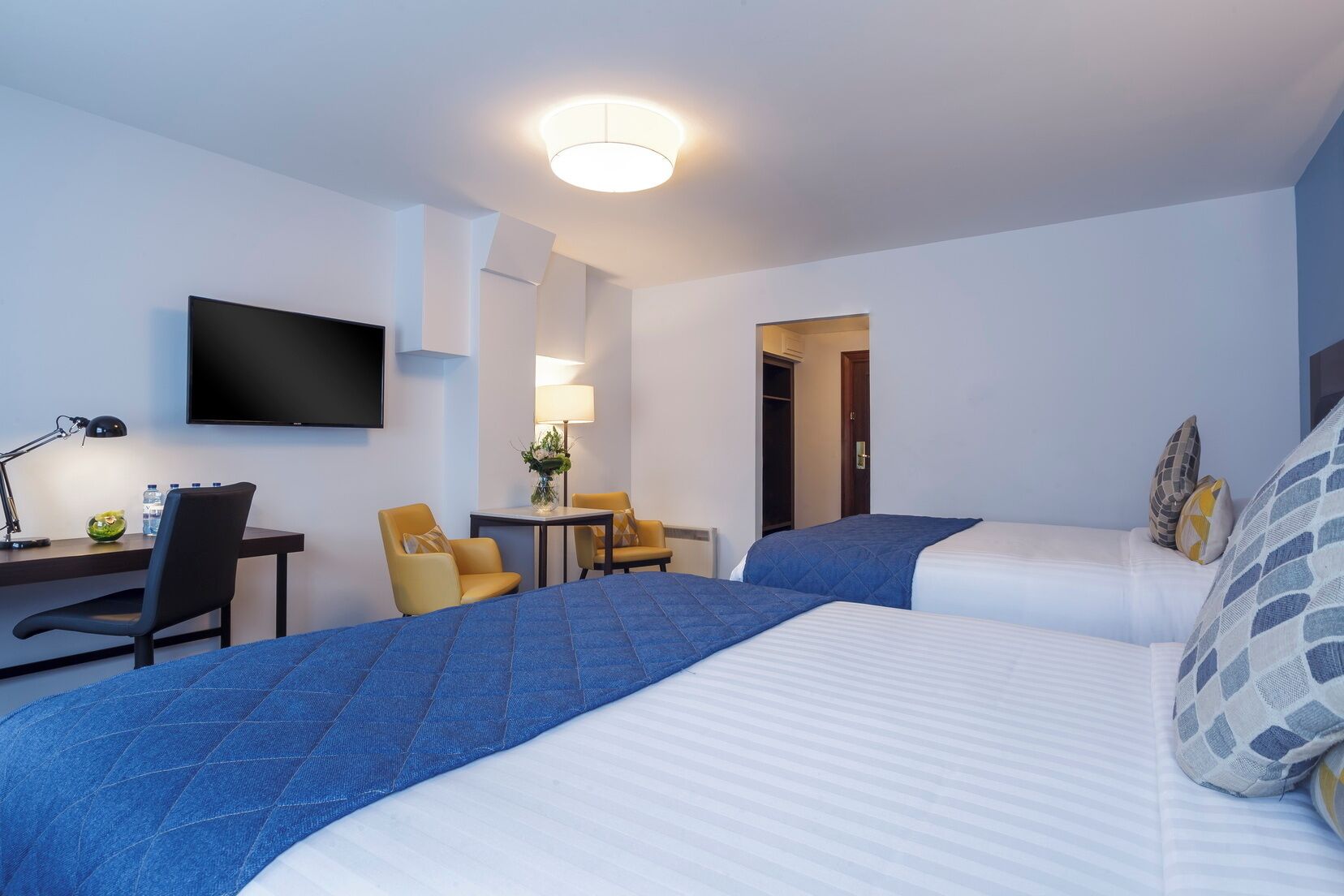 A hotel room with two beds, a desk with a chair, a television, and a small table with a lamp.