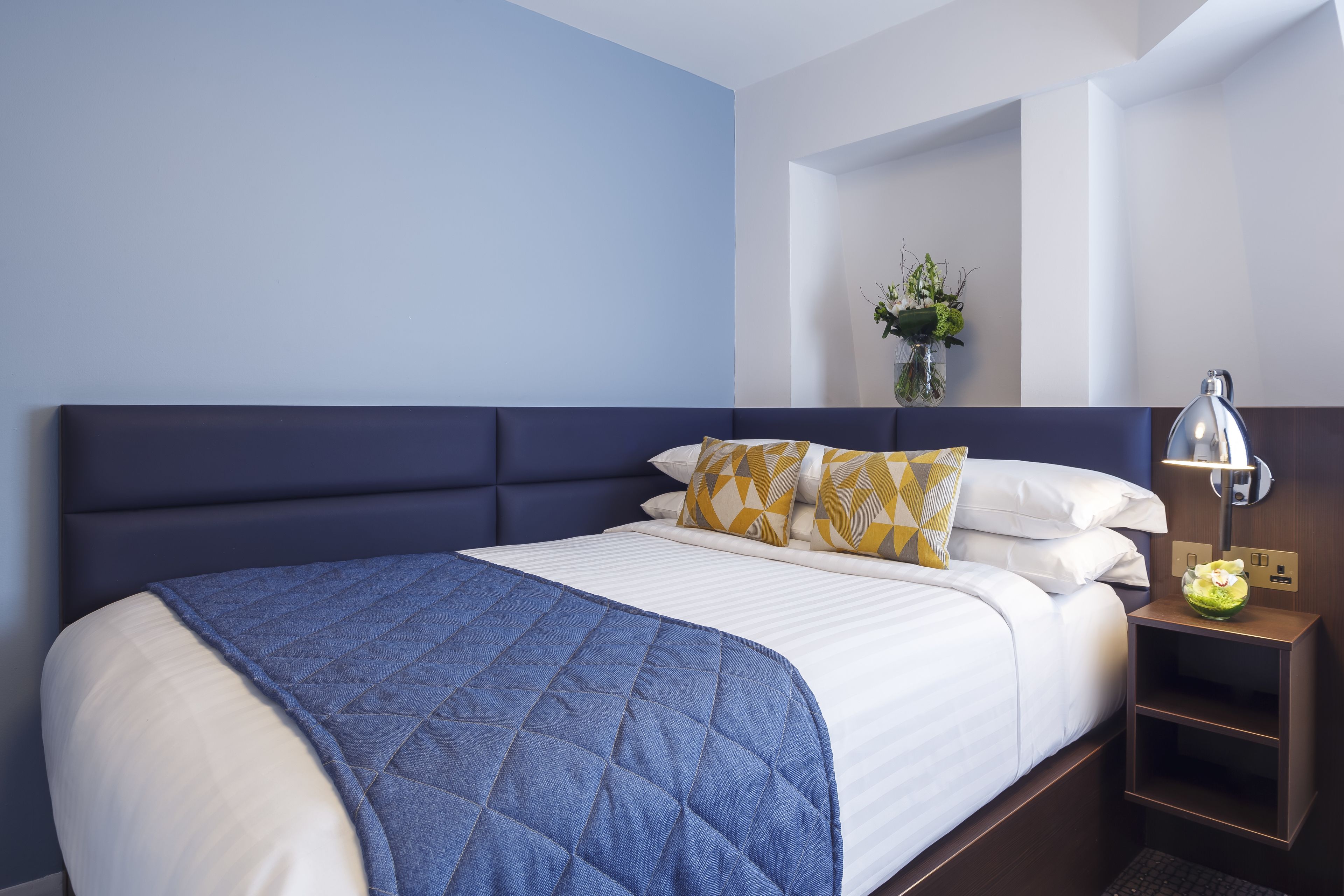 Una cama bien hecha con colcha azul, sábanas blancas y almohadones estampados. Se ve una lámpara de noche y un pequeño jarrón con flores.