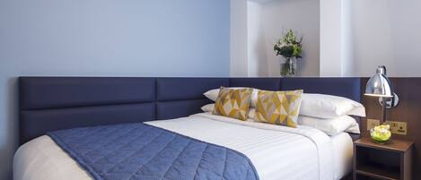 Una cama bien hecha con colcha azul, sábanas blancas y almohadones estampados. Se ve una lámpara de noche y un pequeño jarrón con flores.