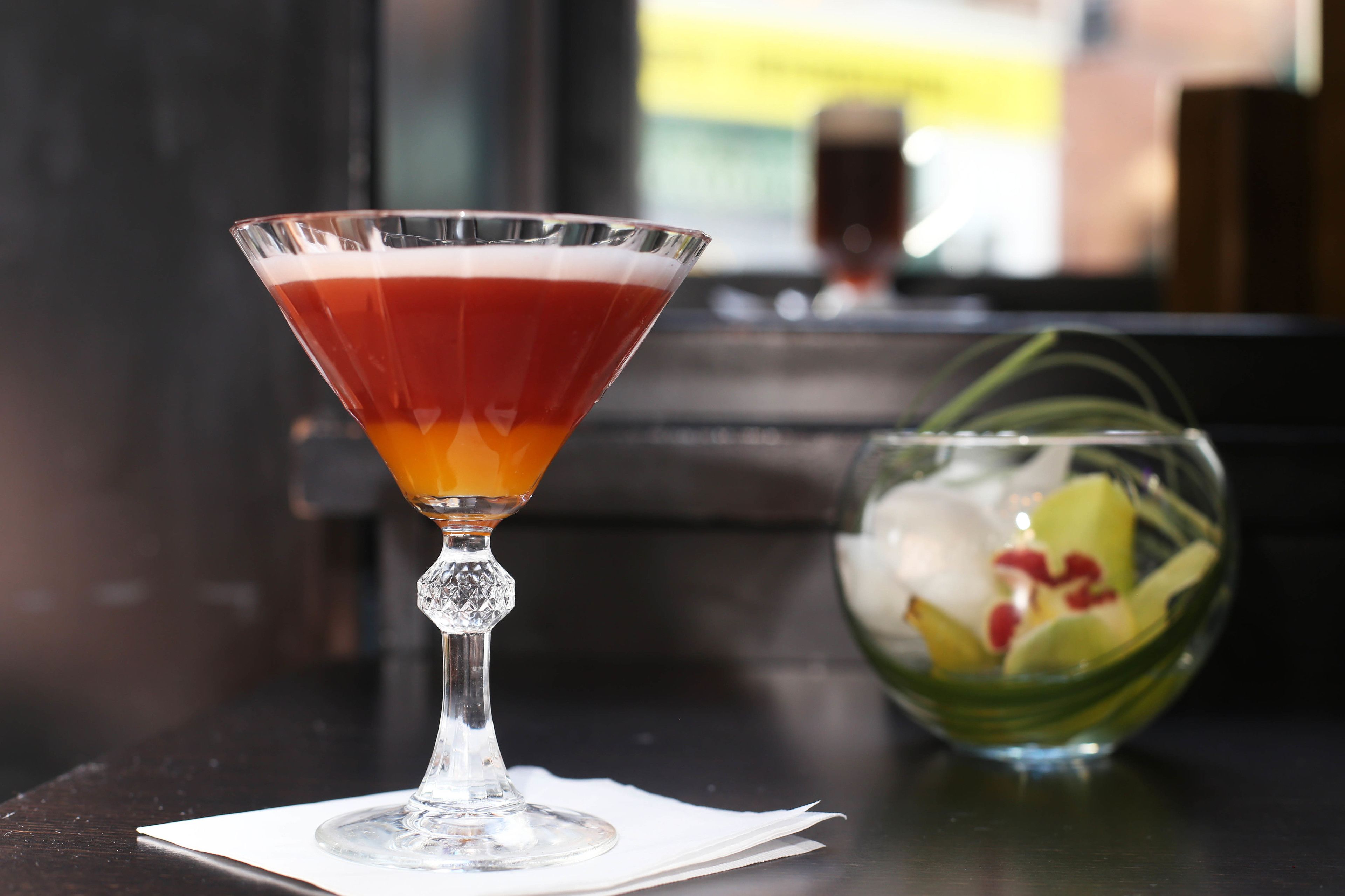 Una copa de martini con una bebida en capas sobre una mesa, junto a un cuenco de fruta y una servilleta blanca.
