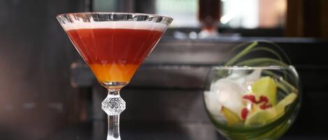 A martini glass with a layered drink on a table, next to a bowl of fruit and a white napkin.