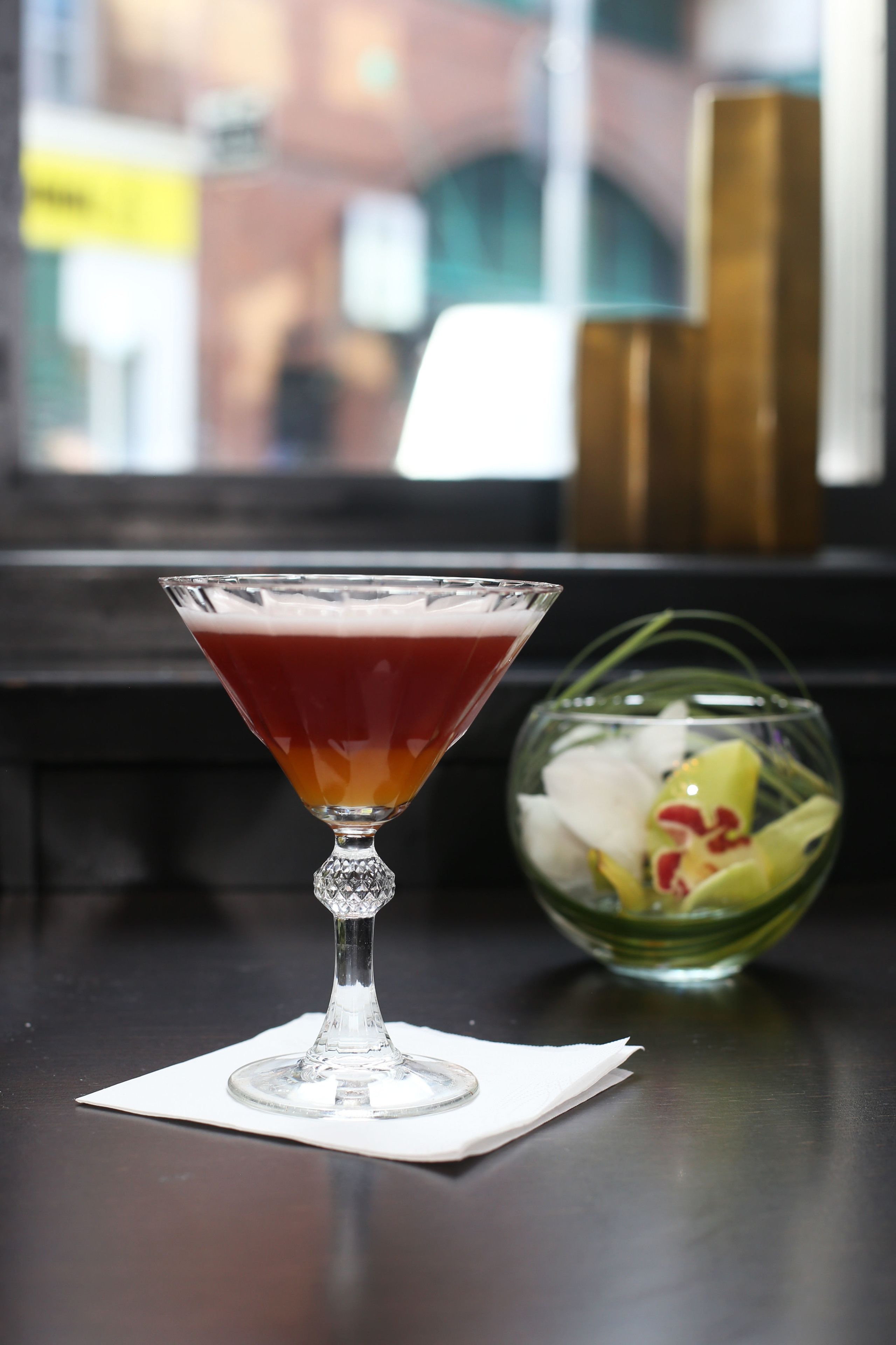 Una copa de martini con una bebida en capas sobre una mesa, junto a un florero con flores.
