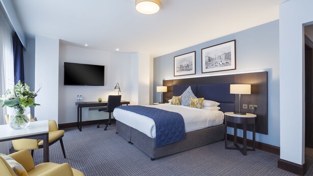 A modern hotel room with a large bed, a desk, a chair, a TV, and framed pictures on the wall.