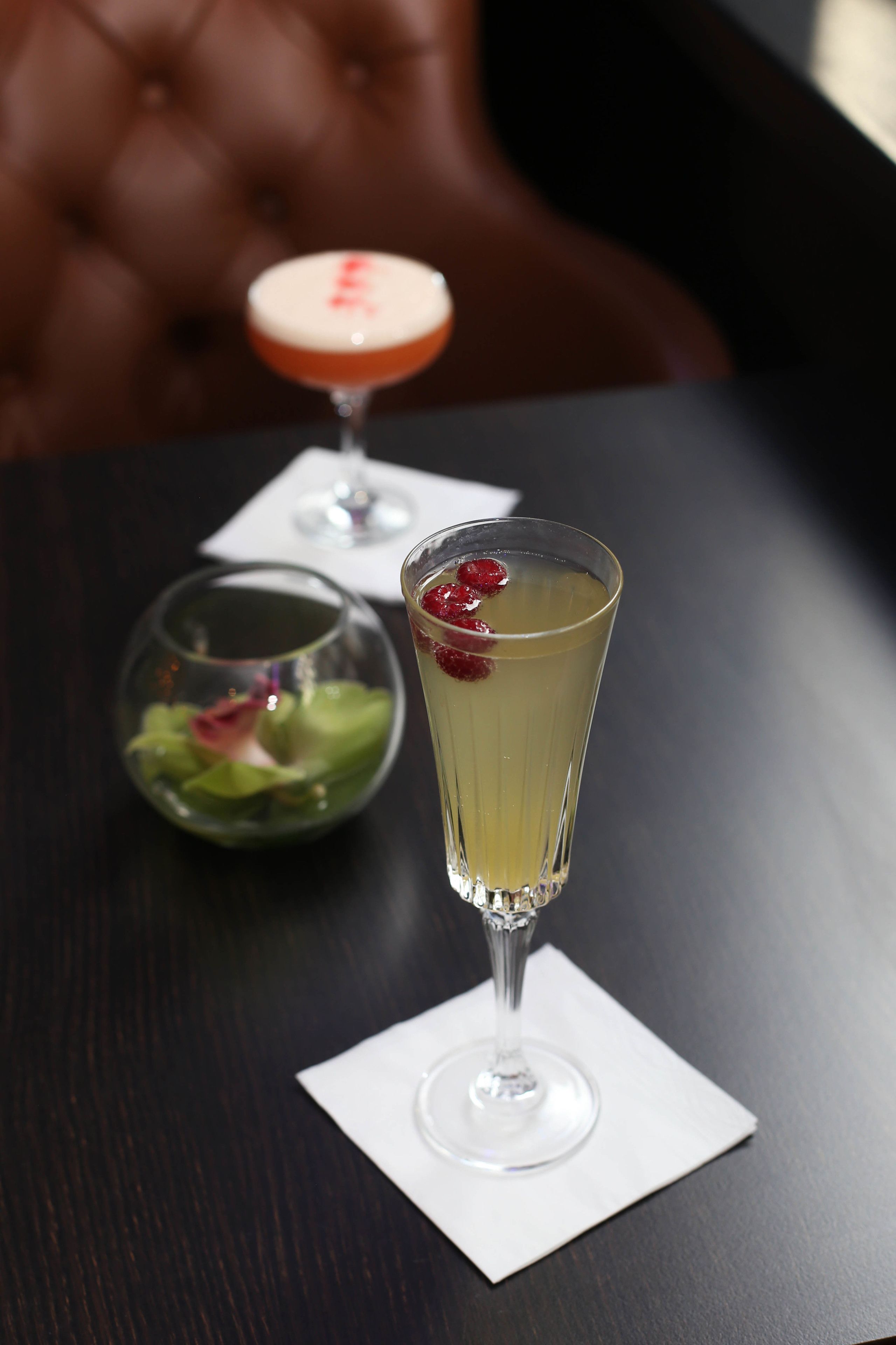 A table with a tall glass of a yellow beverage garnished with berries, a smaller glass with a red rim, and a bowl of green salad.