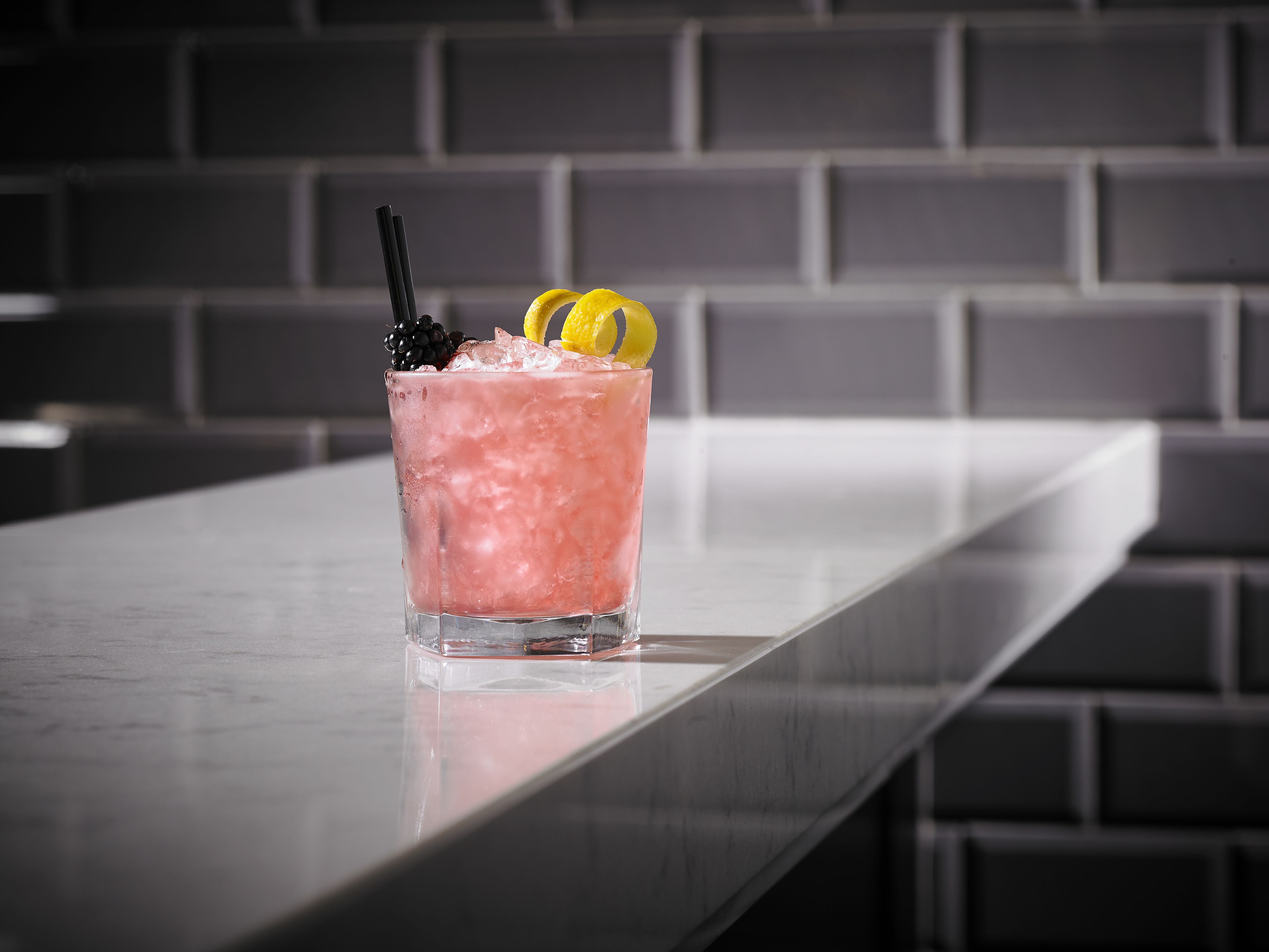A glass of pink beverage with blackberries and lemon slices on a bar counter.
