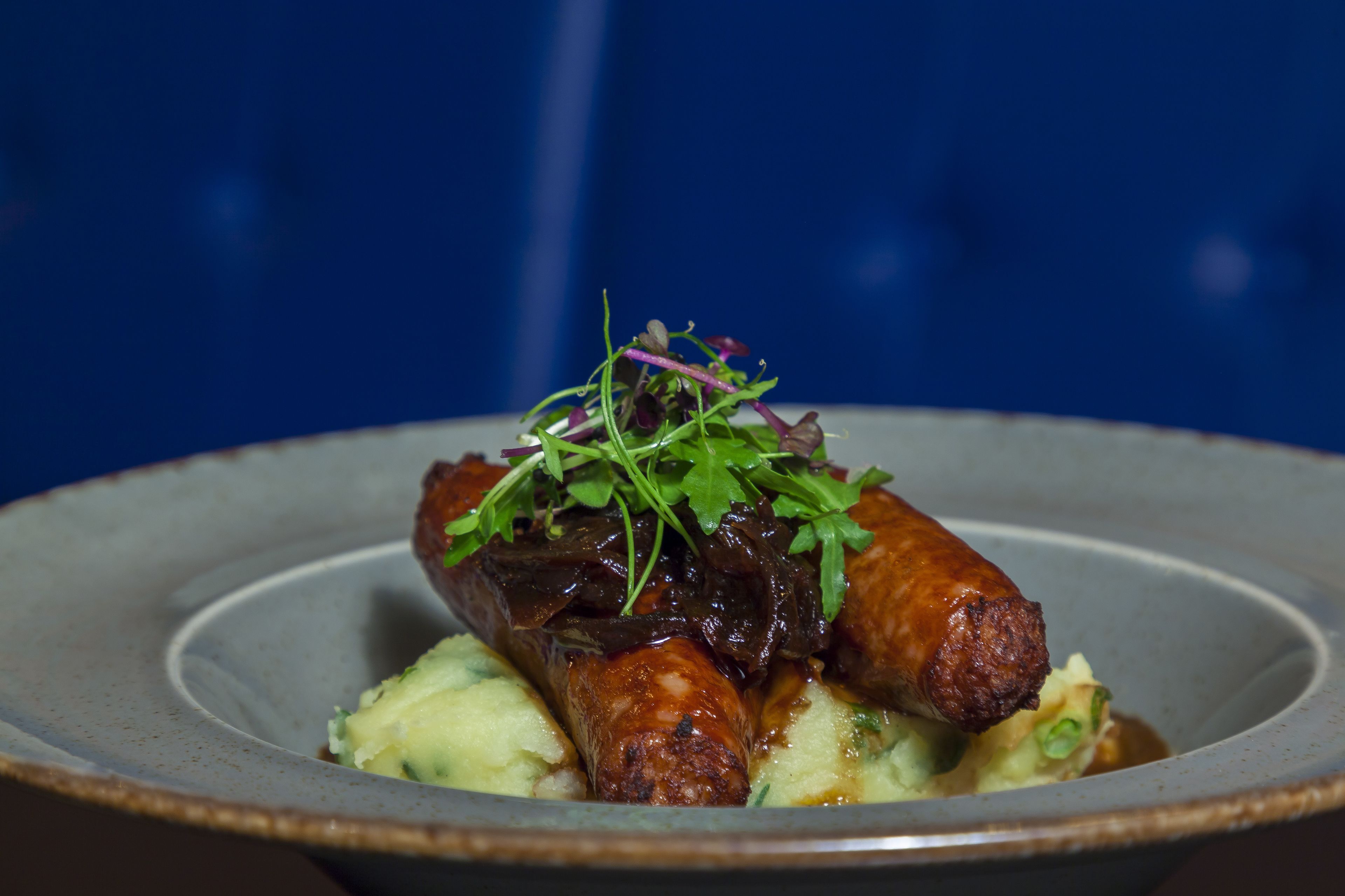 Un plato con salchicha, puré de patatas y guarnición de vegetales verdes.