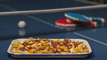 A table with a dish of fries topped with cheese, bacon, and sauce. A ping pong table and paddles are in the background.