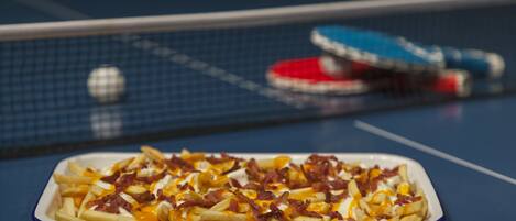 Una mesa con un plato de papas fritas cubiertas de queso, tocino y salsa. En el fondo, una mesa de ping pong y paletas.