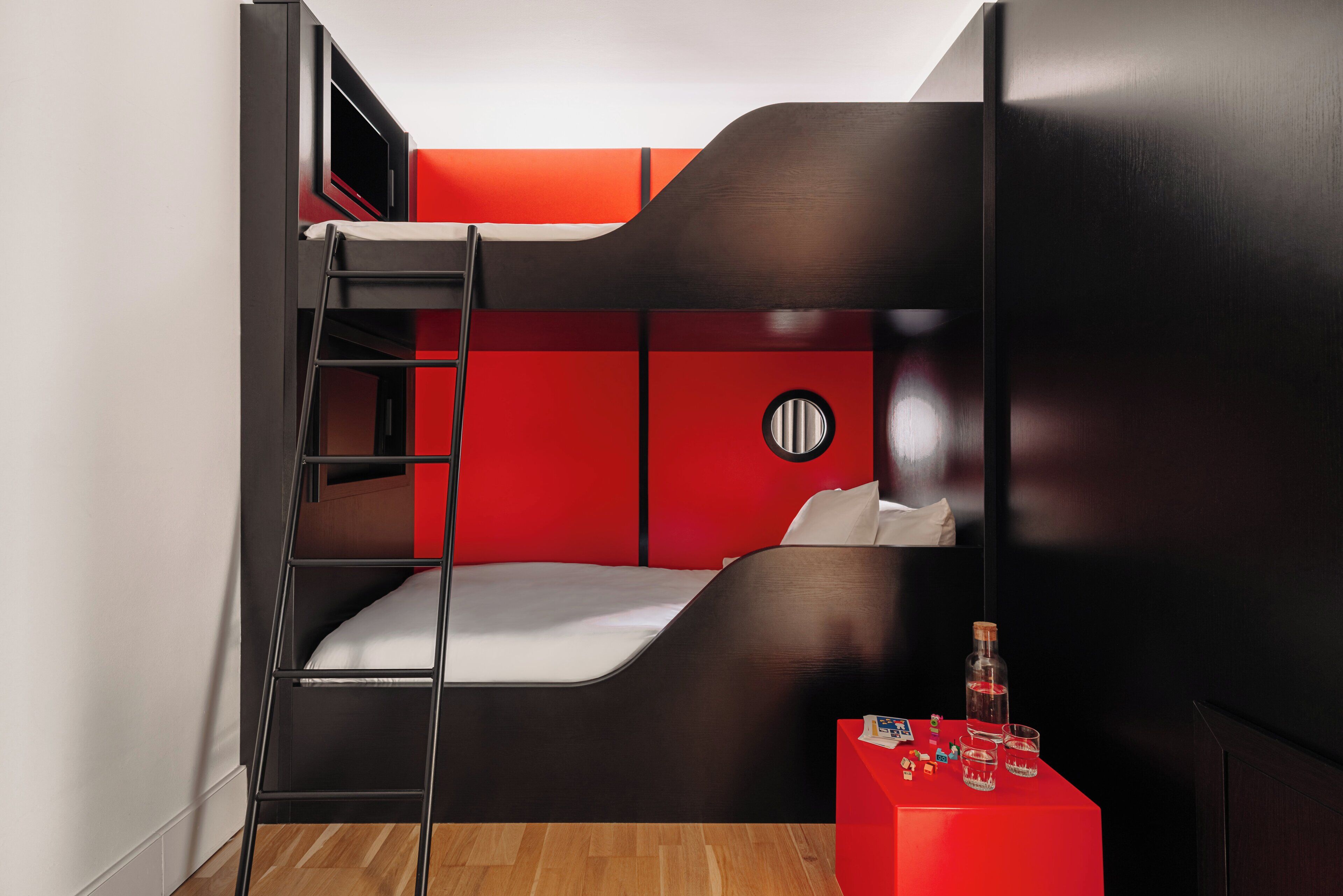 A bunk bed room with a ladder, a red table with bottles, and a round wall decoration.