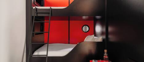 Habitación con literas, una escalera, una mesa roja con botellas y un adorno circular en la pared.