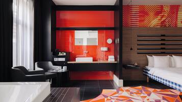 Een moderne hotelkamer met een groot bed, een zithoek met een stoel en tafel, een badkamer met een rode accentmuur en een tapijt met een geometrisch patroon.