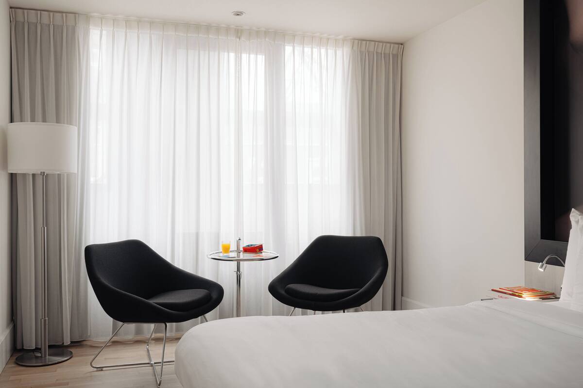 A hotel room with a bed, two black chairs, a small table with a drink and a red object, a floor lamp, and a large window with sheer curtains.