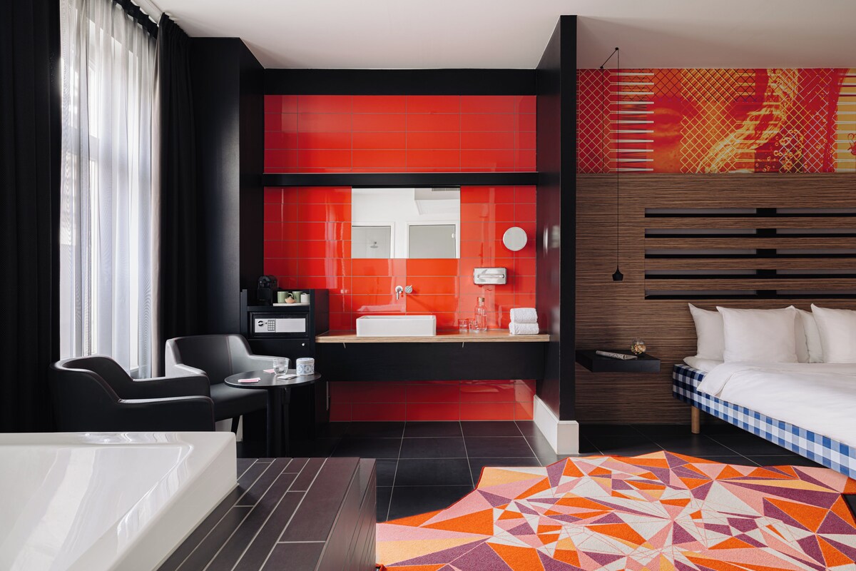 Une chambre d’hôtel moderne dotée d’un grand lit, d’un coin salon séparé, d’une salle de bain avec un mur d’accent rouge et d’un tapis à motifs géométriques.