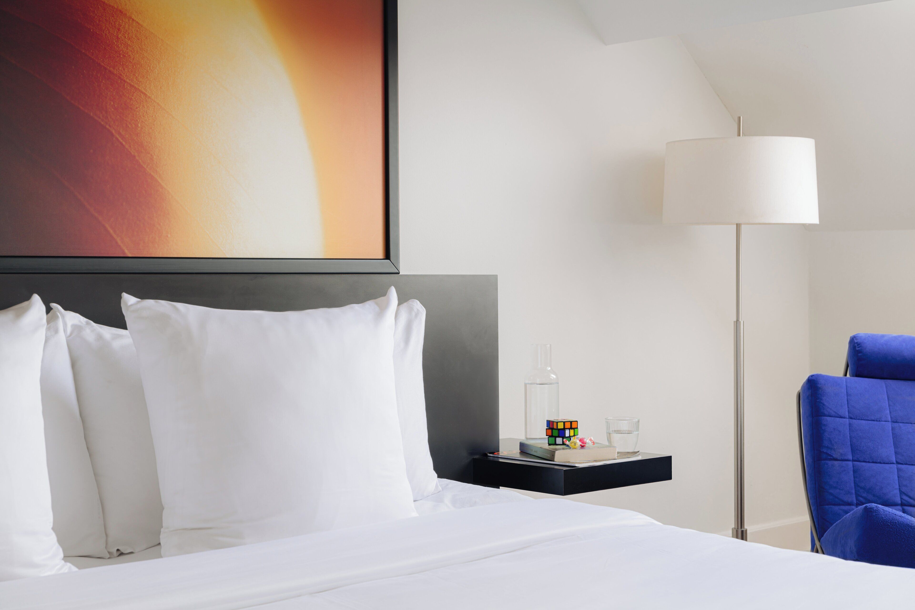 A hotel room with a neatly made bed, a bedside table with a lamp, a chair, and a wall art piece.