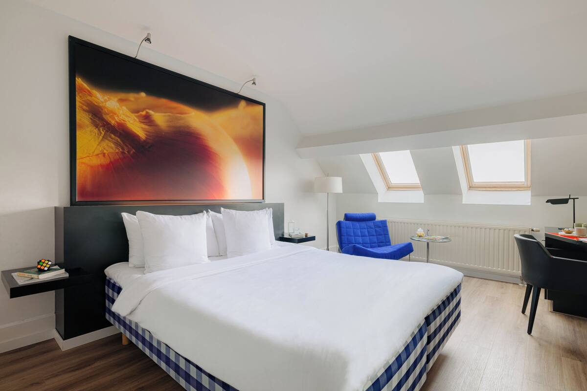 A modern hotel room with a large bed, a desk, a chair, and a painting on the wall.
