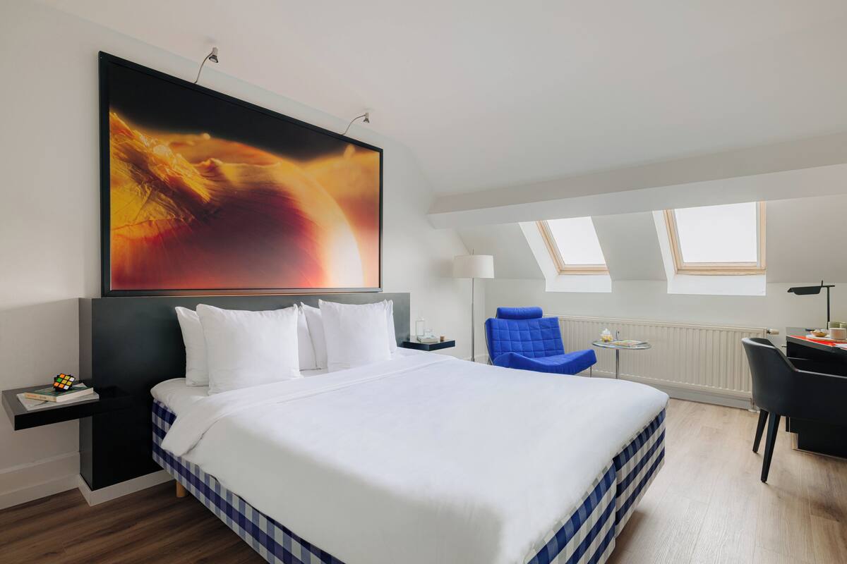 A modern hotel room with a large bed, a desk, a chair, and a painting on the wall.