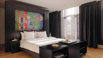 Une chambre d’hôtel moderne avec un grand lit, une table de chevet noire, une table d’appoint, une console noire, une peinture abstraite aux couleurs vives et une grande fenêtre avec des rideaux noirs.