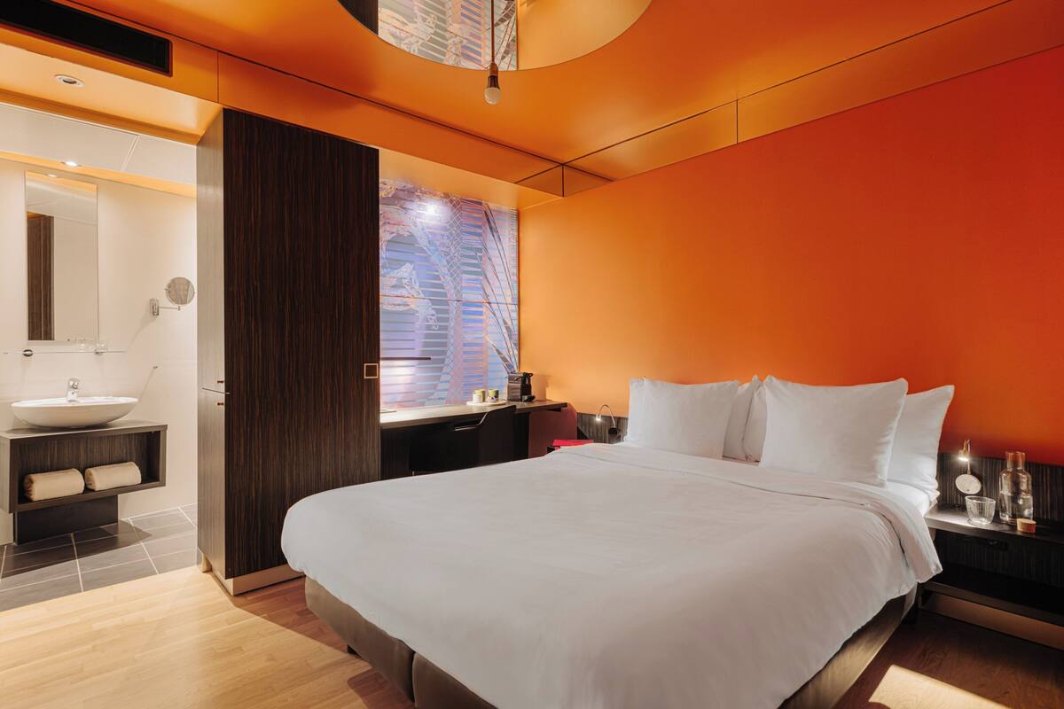 A modern hotel room with a large bed, a bathroom with a sink and mirror, and a view of a building through a window.