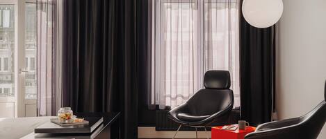 Una habitación de hotel moderna con un ventanal grande, un escritorio, dos sillas negras, una mesita de noche roja y una lámpara colgante.