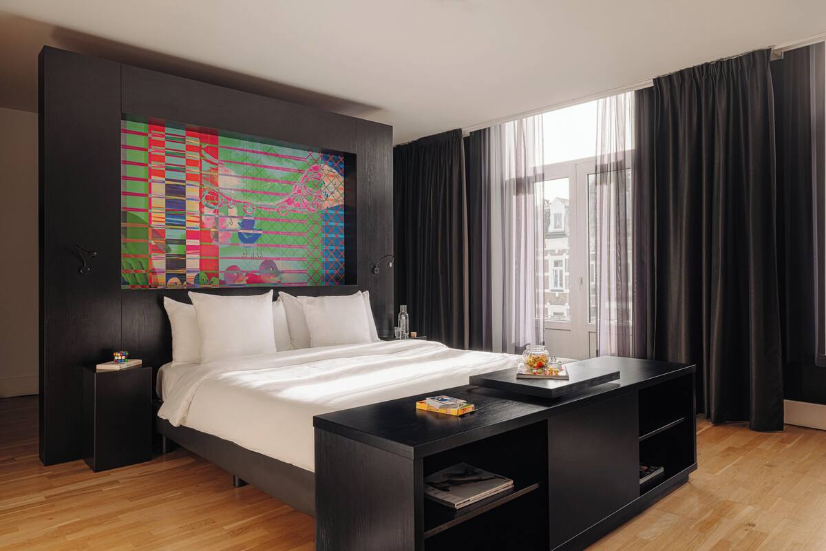 A modern hotel room with a large bed, a black nightstand, a bedside table, a black console table, a colorful abstract painting, and a large window with black curtains.
