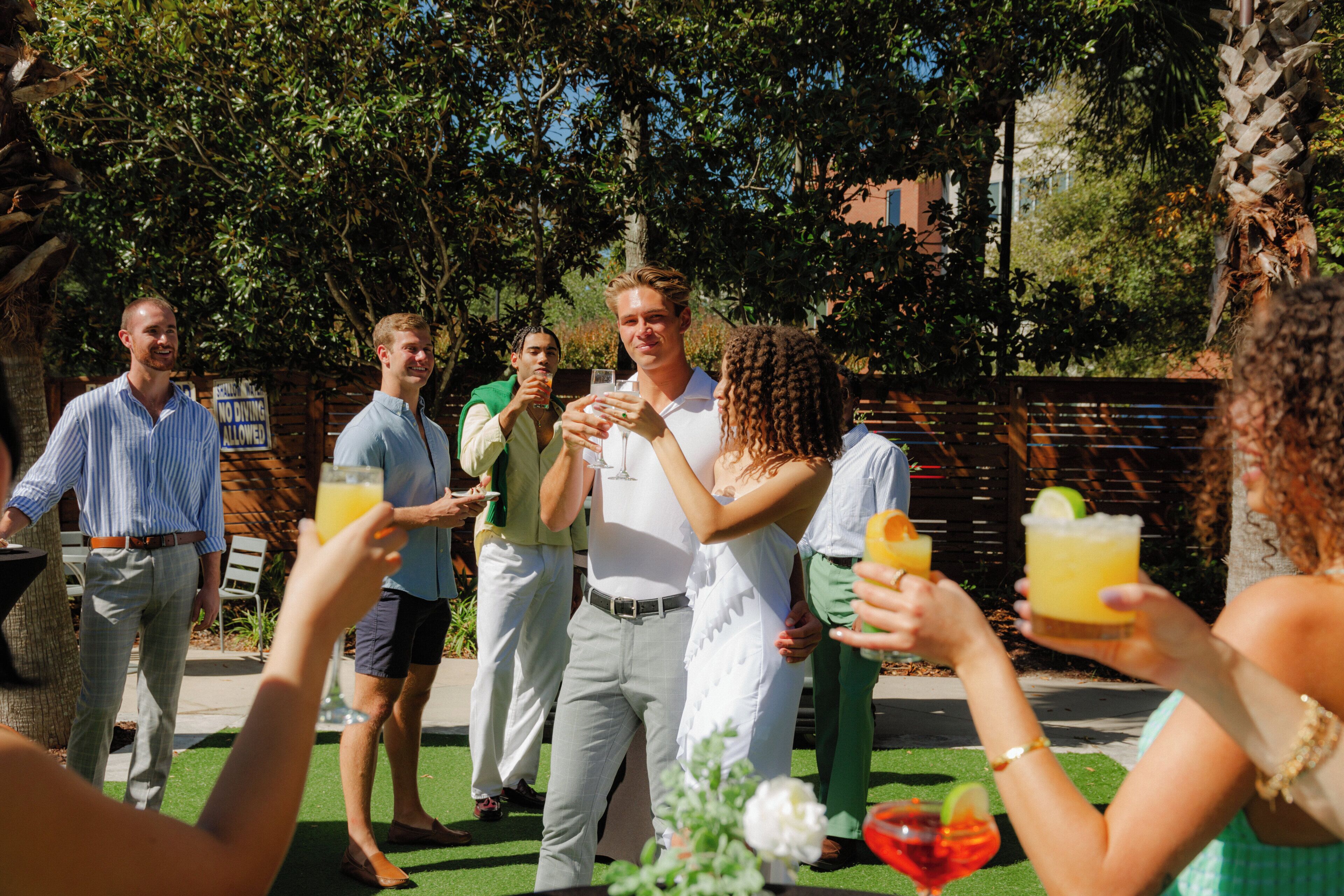 A group of people toasting with drinks in an outdoor setting with greenery and a fence in the background.