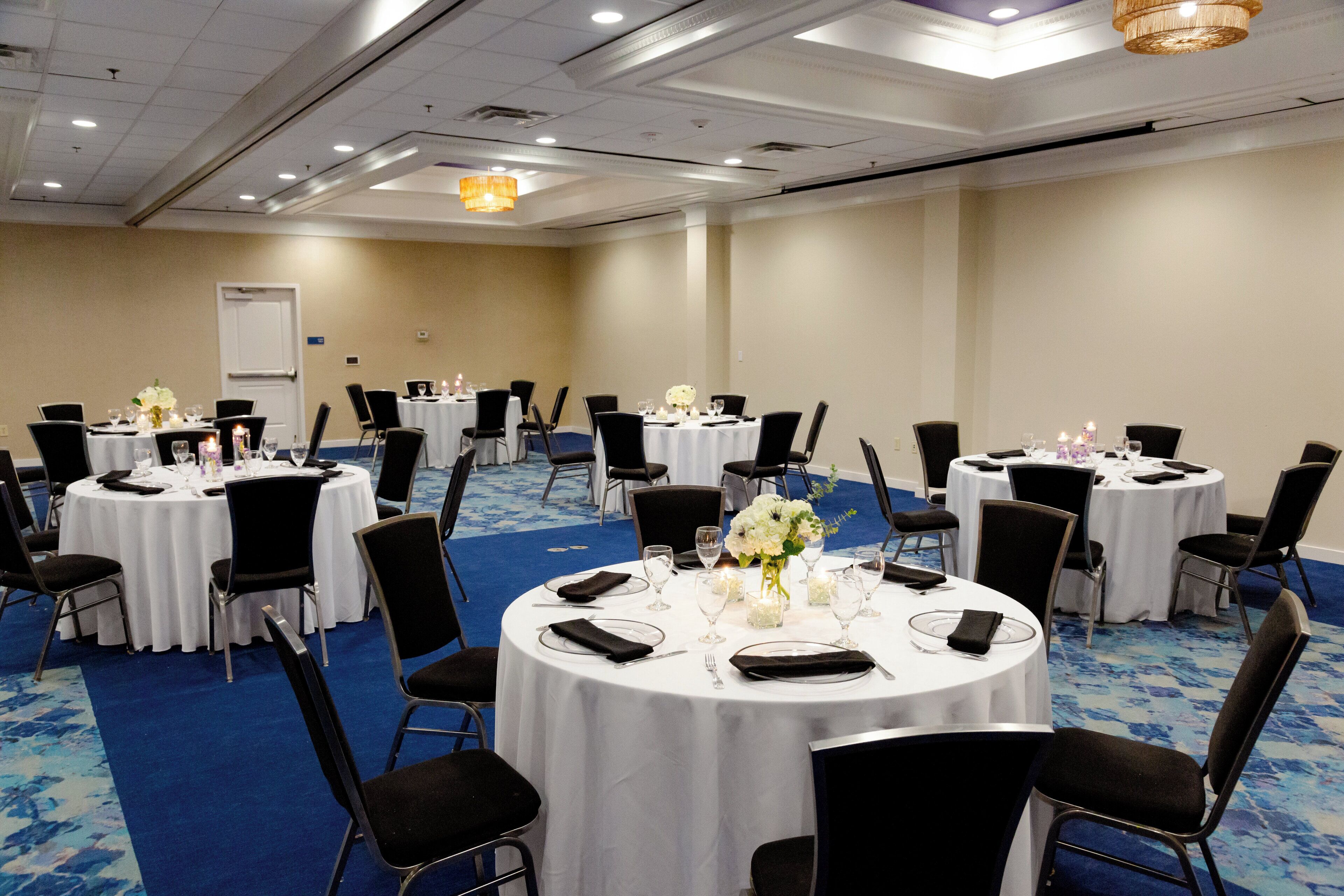 Une salle de conférence équipée de plusieurs tables rondes, recouvertes de nappes blanches, avec des chaises noires et agrémentées de compositions florales.