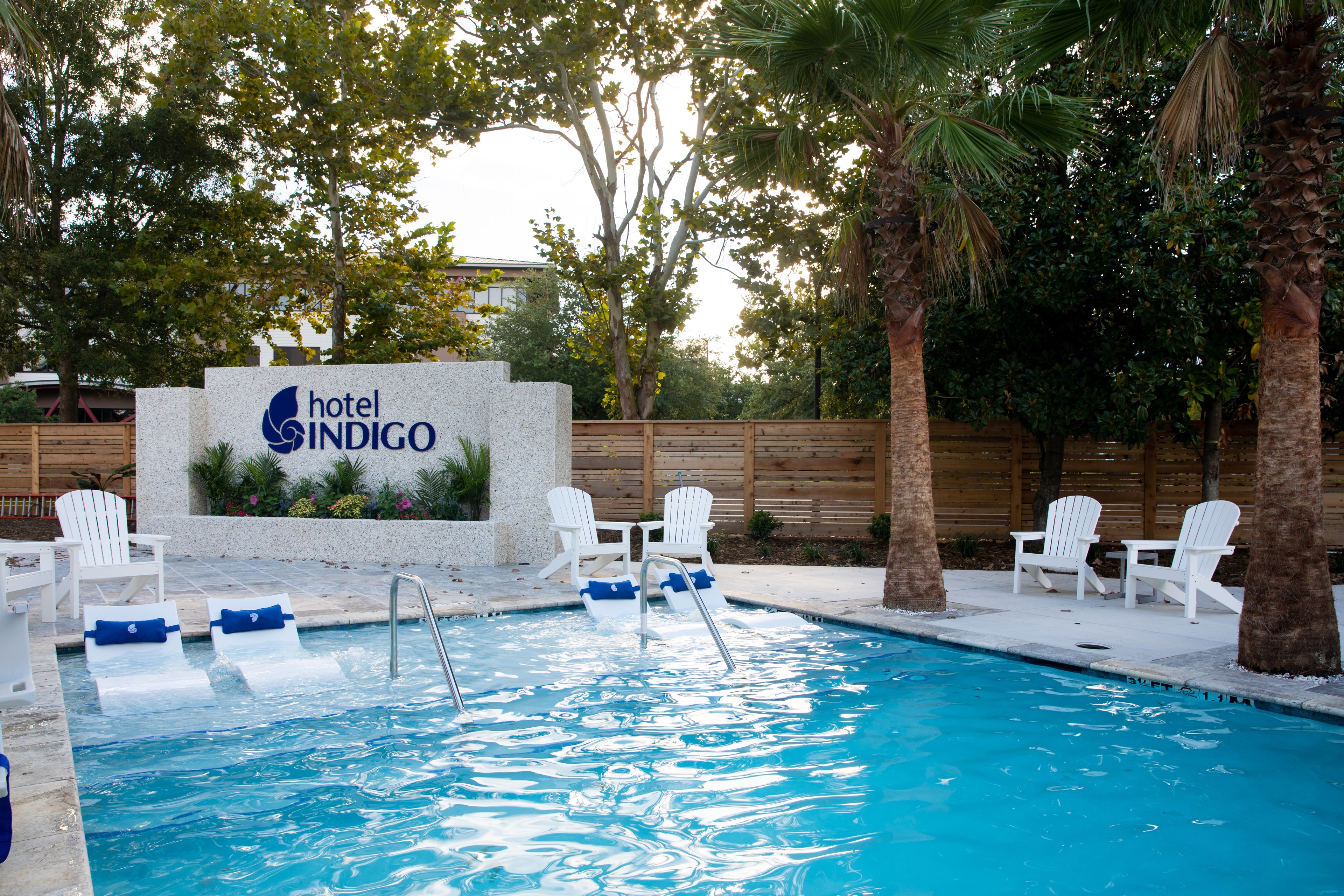 Zwembadgebied van een hotel, met een bord waarop 'hotel INDIGO' staat, omringd door witte stoelen en palmbomen.