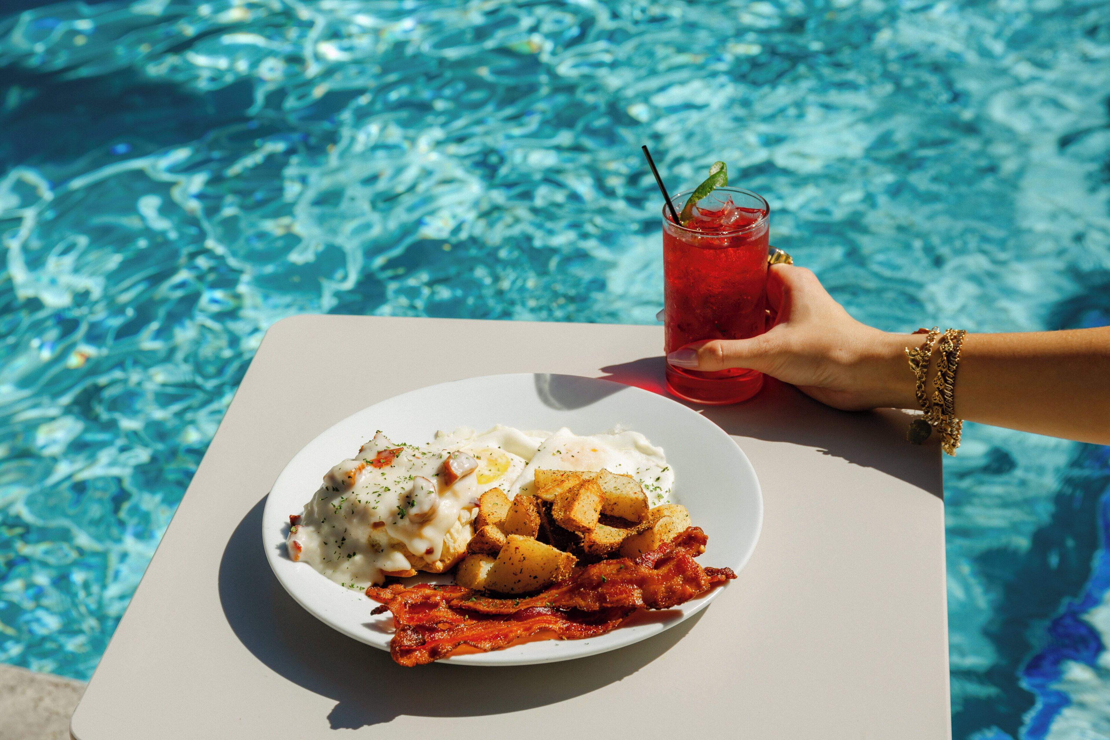 A plate of food with eggs, potatoes, and bacon, and a glass of red drink with a straw, by a pool.