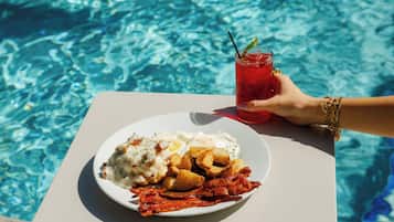 A plate of food with eggs, potatoes, and bacon, and a glass of red drink with a straw, by a pool.