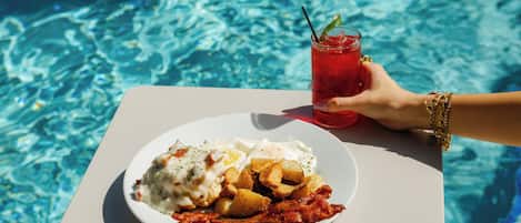 A plate of food with eggs, potatoes, and bacon, and a glass of red drink with a straw, by a pool.