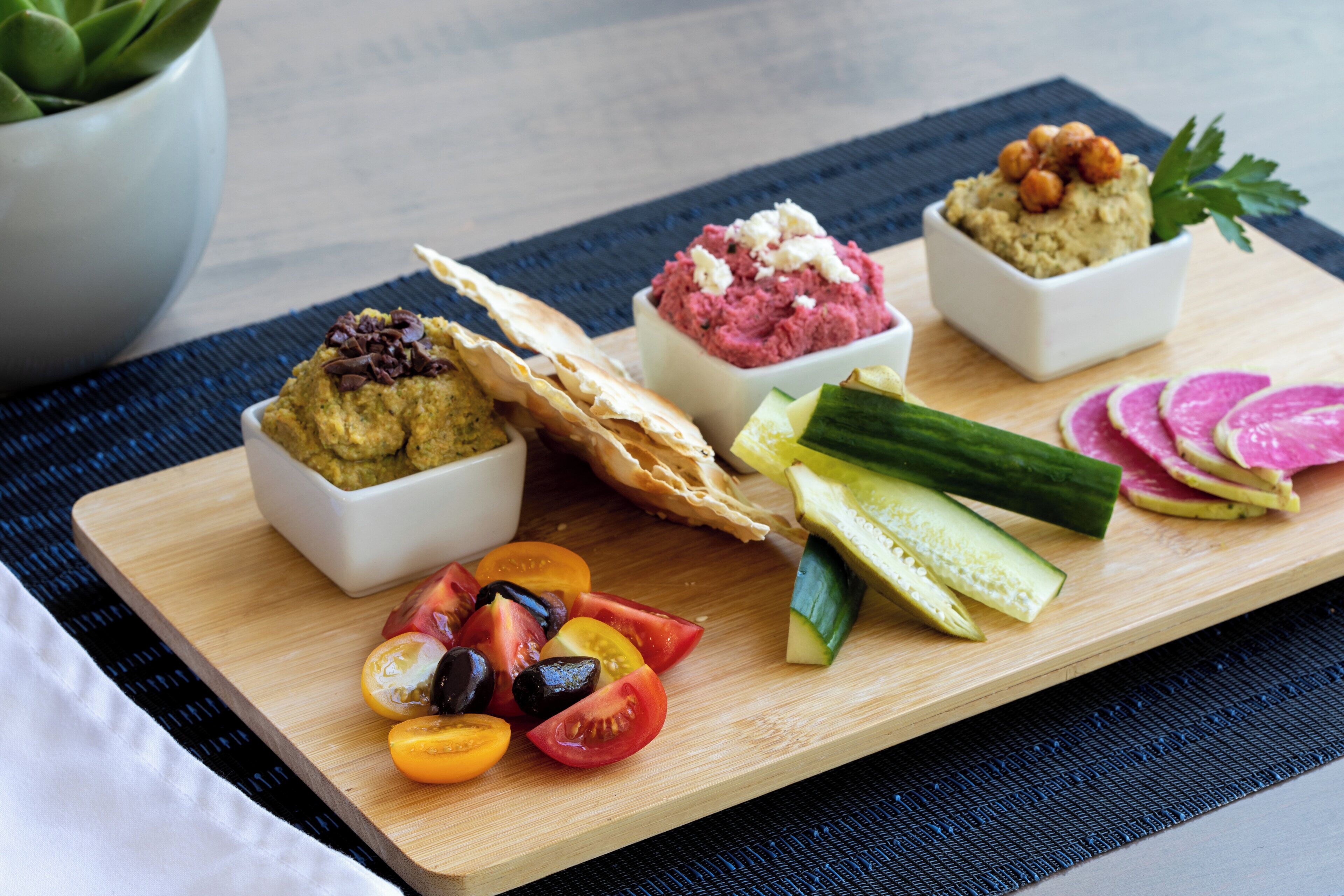 A wooden board with various food items including hummus, a flatbread, and sliced vegetables.