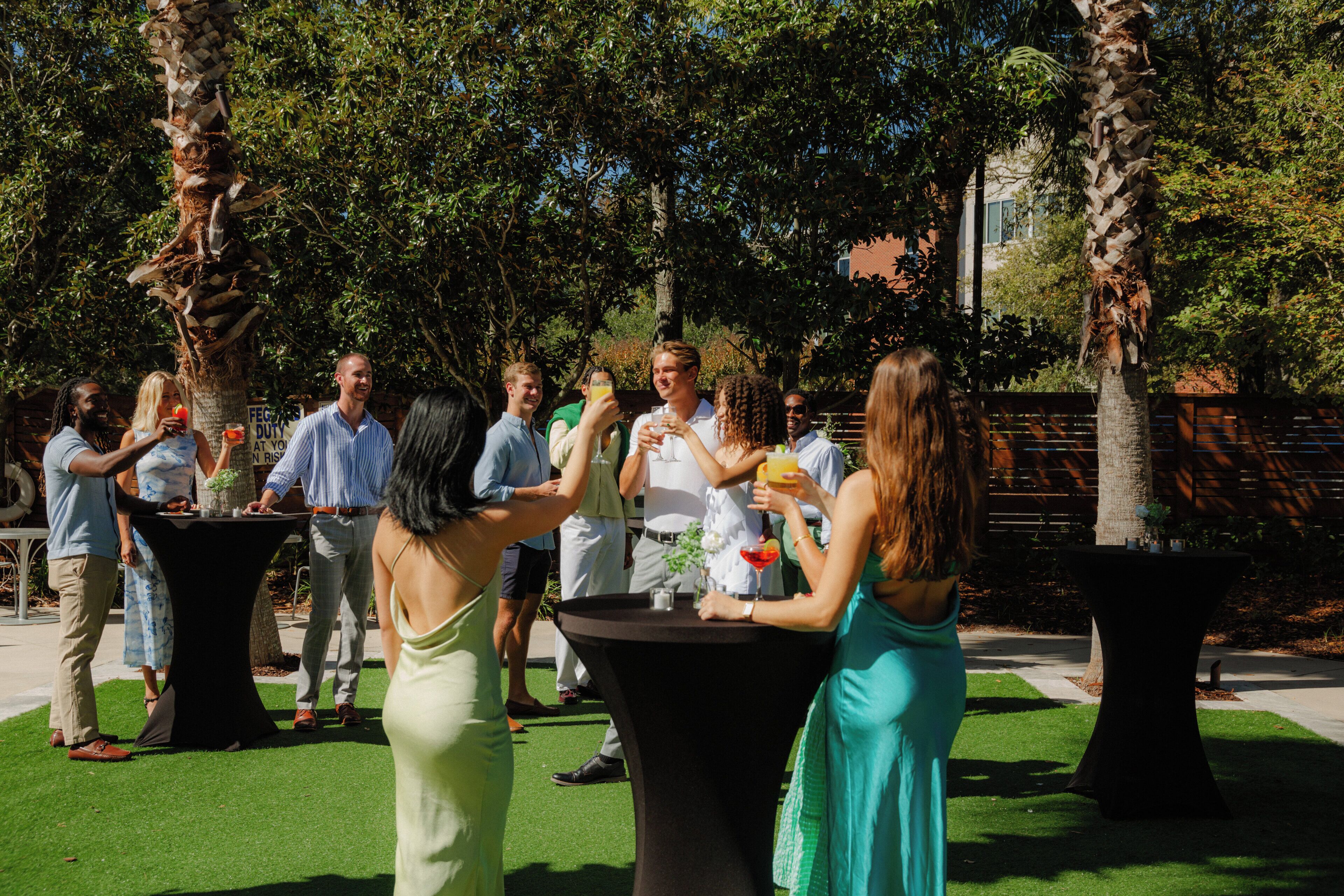A group of people standing around tables in an outdoor setting with palm trees and a brick wall in the background.