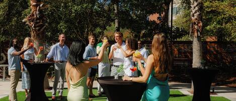 A group of people standing around tables in an outdoor setting with palm trees and a brick wall in the background.