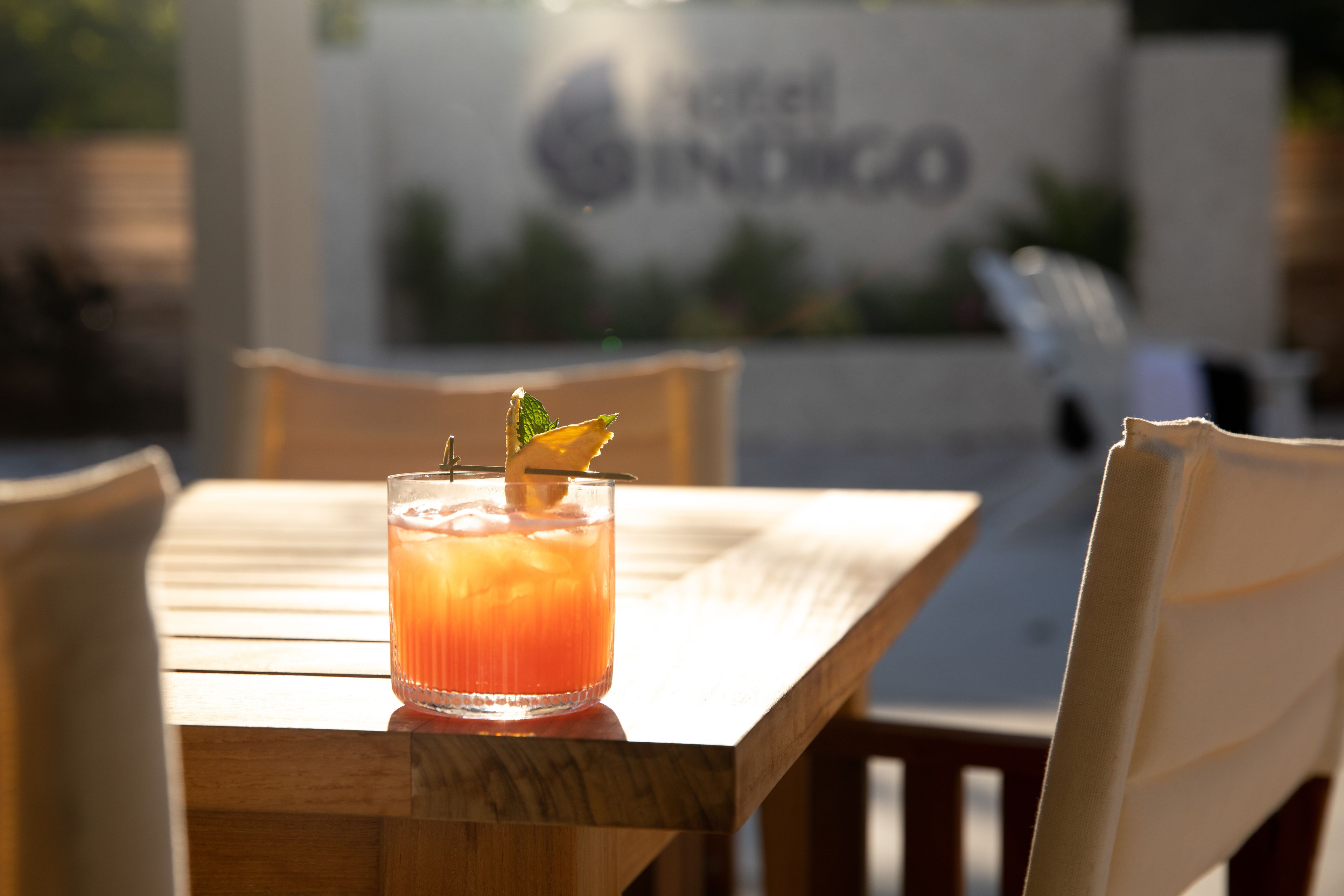 A wooden table with a glass of orange drink garnished with a mint leaf, set against a blurred background with a sign reading "Hotel Spinico".
