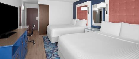 A hotel room with a large bed, a smaller bed, a desk with a TV, and a bathroom with a mirror and towel rack.