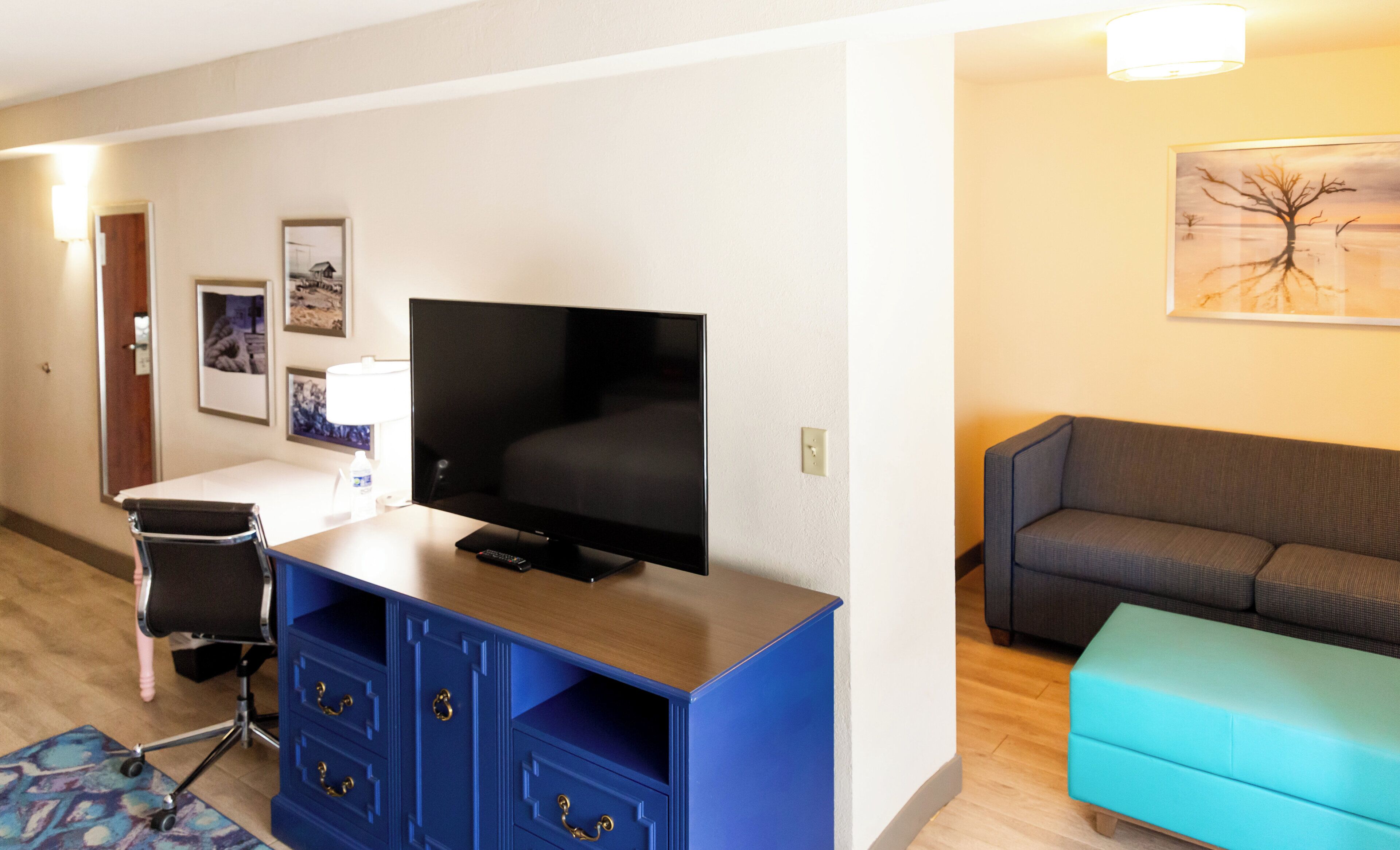 A hotel room with a flat-screen TV, a blue cabinet with drawers, a grey sofa, a teal ottoman, and a framed picture on the wall.