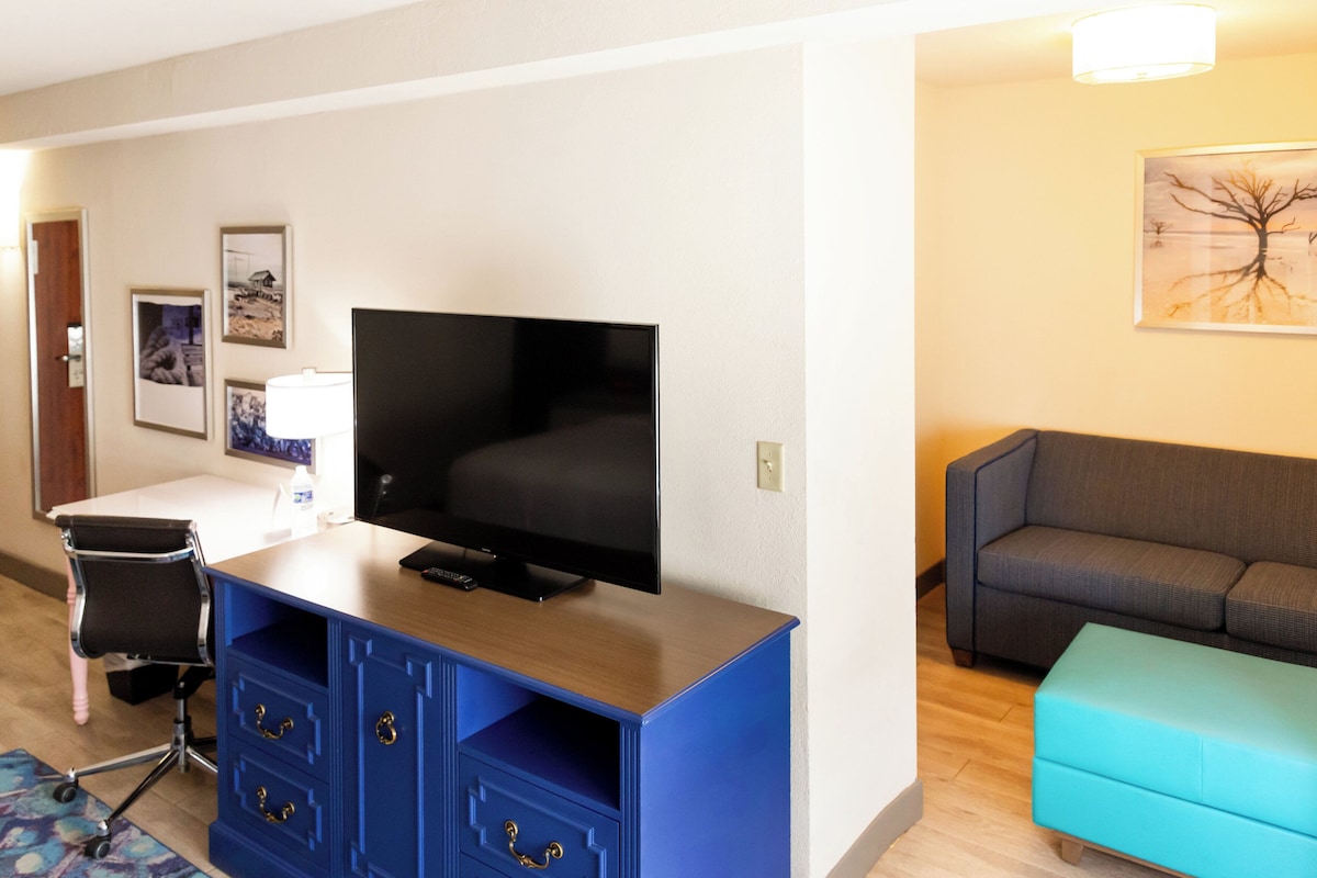 A hotel room with a flat-screen TV, a blue cabinet with drawers, a grey sofa, a teal ottoman, and a framed picture on the wall.