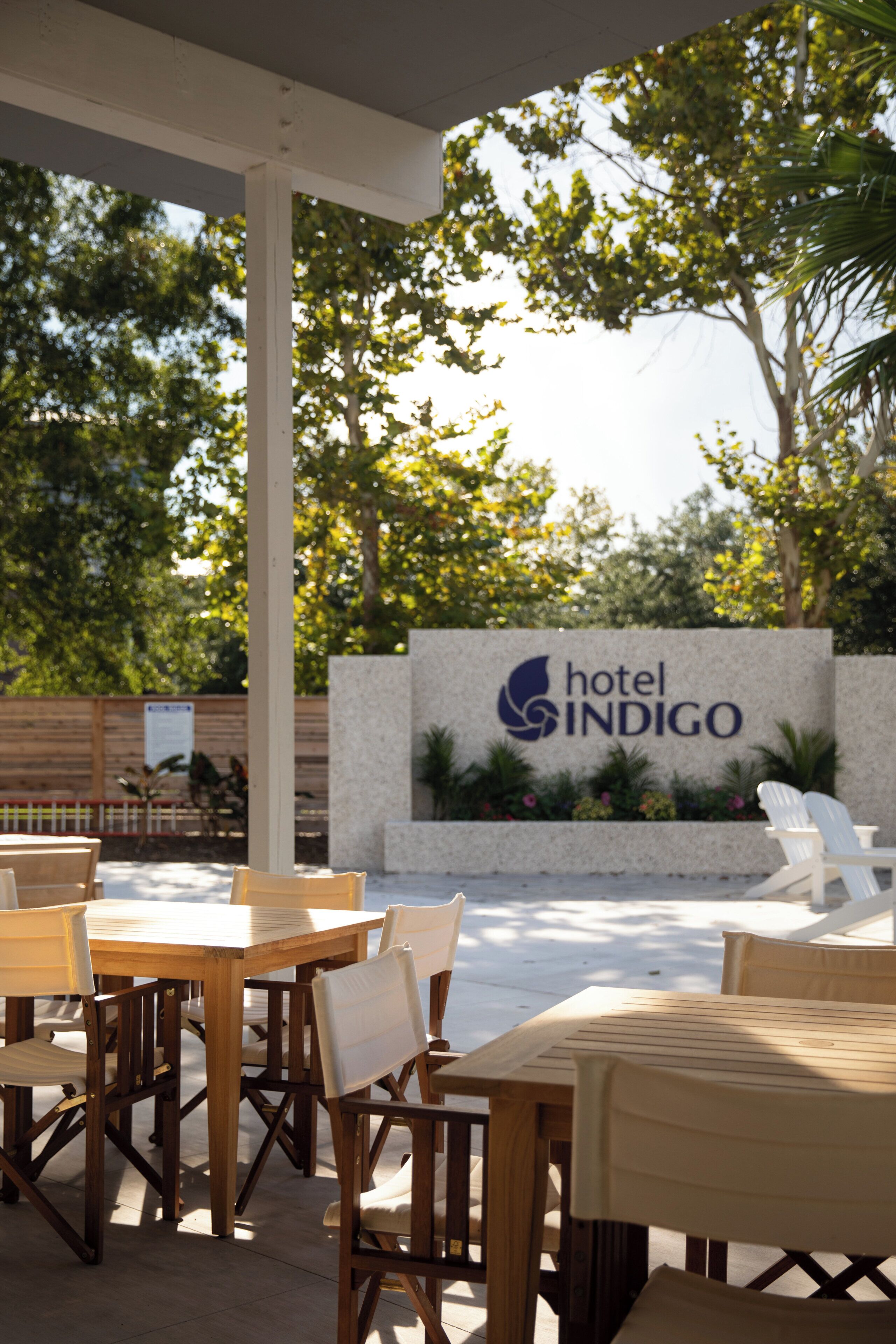 An outdoor seating area with wooden tables and chairs, a stone wall with the 'Hotel Indigo' sign, and surrounding greenery.
