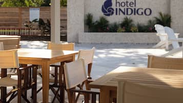 An outdoor seating area with wooden tables and chairs, a stone wall with the 'Hotel Indigo' sign, and surrounding greenery.